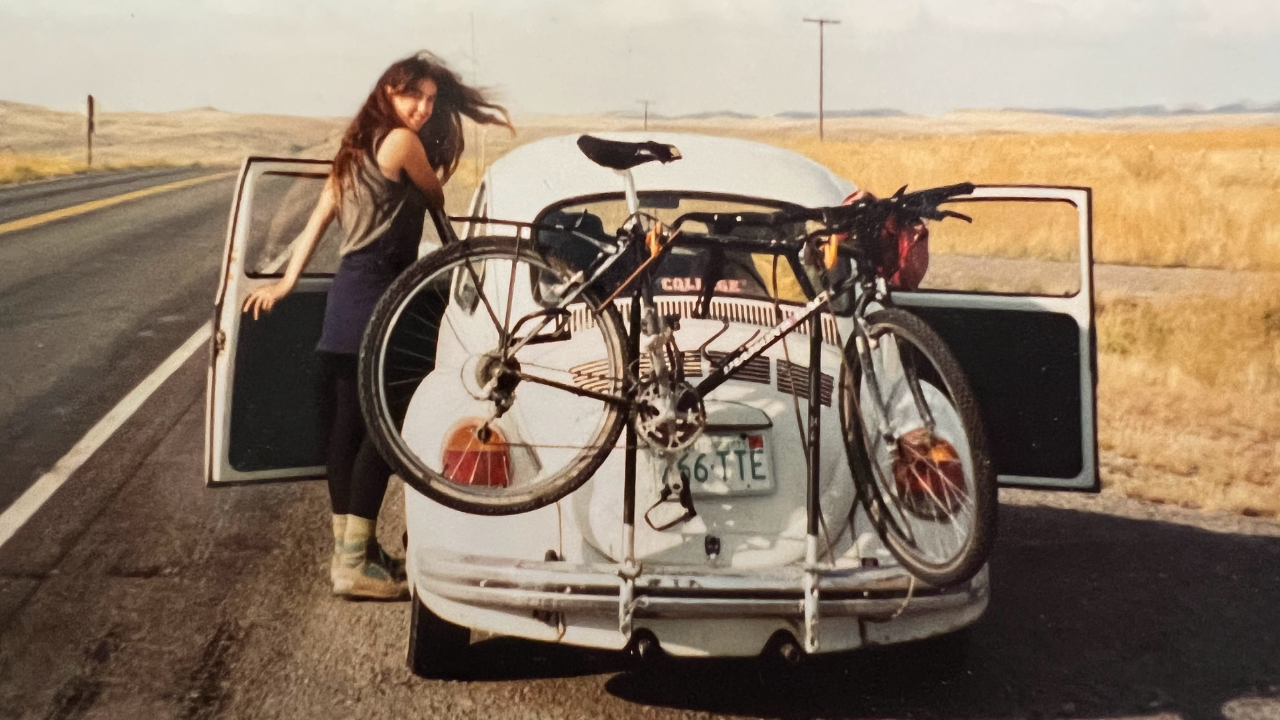 Chantal, owner of Emerging Women, standing by the side of the road in the doorway of a vehicle. The car is a light blue bug with a bicycle attached to the back of it.